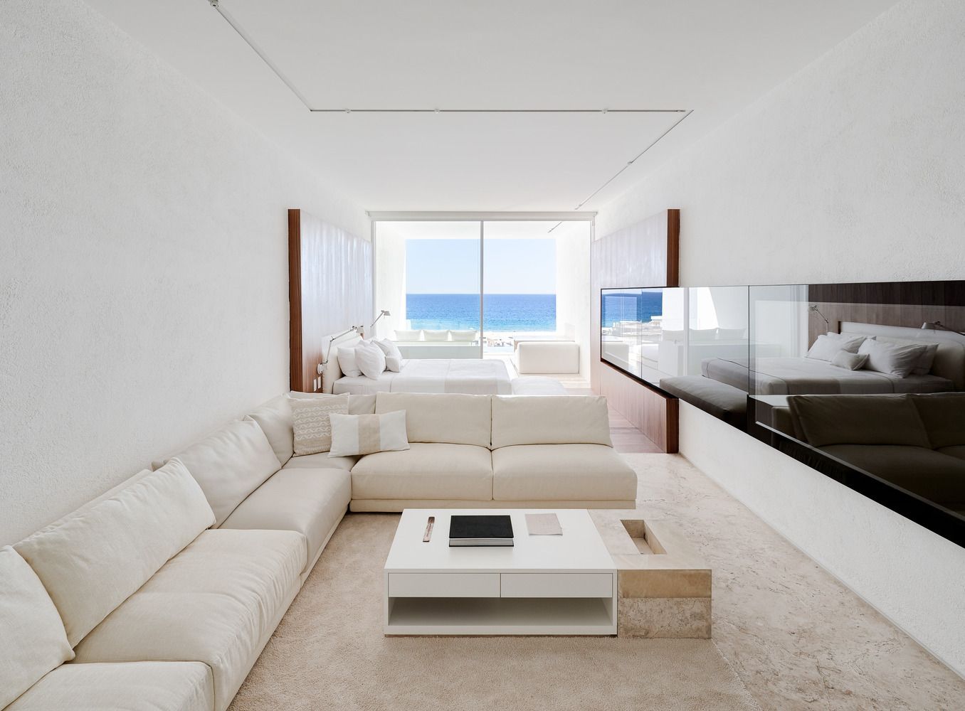 Luminoso salón minimalista con sofás seccionales blancos, mesa de centro y vistas al océano a través de grandes ventanales.