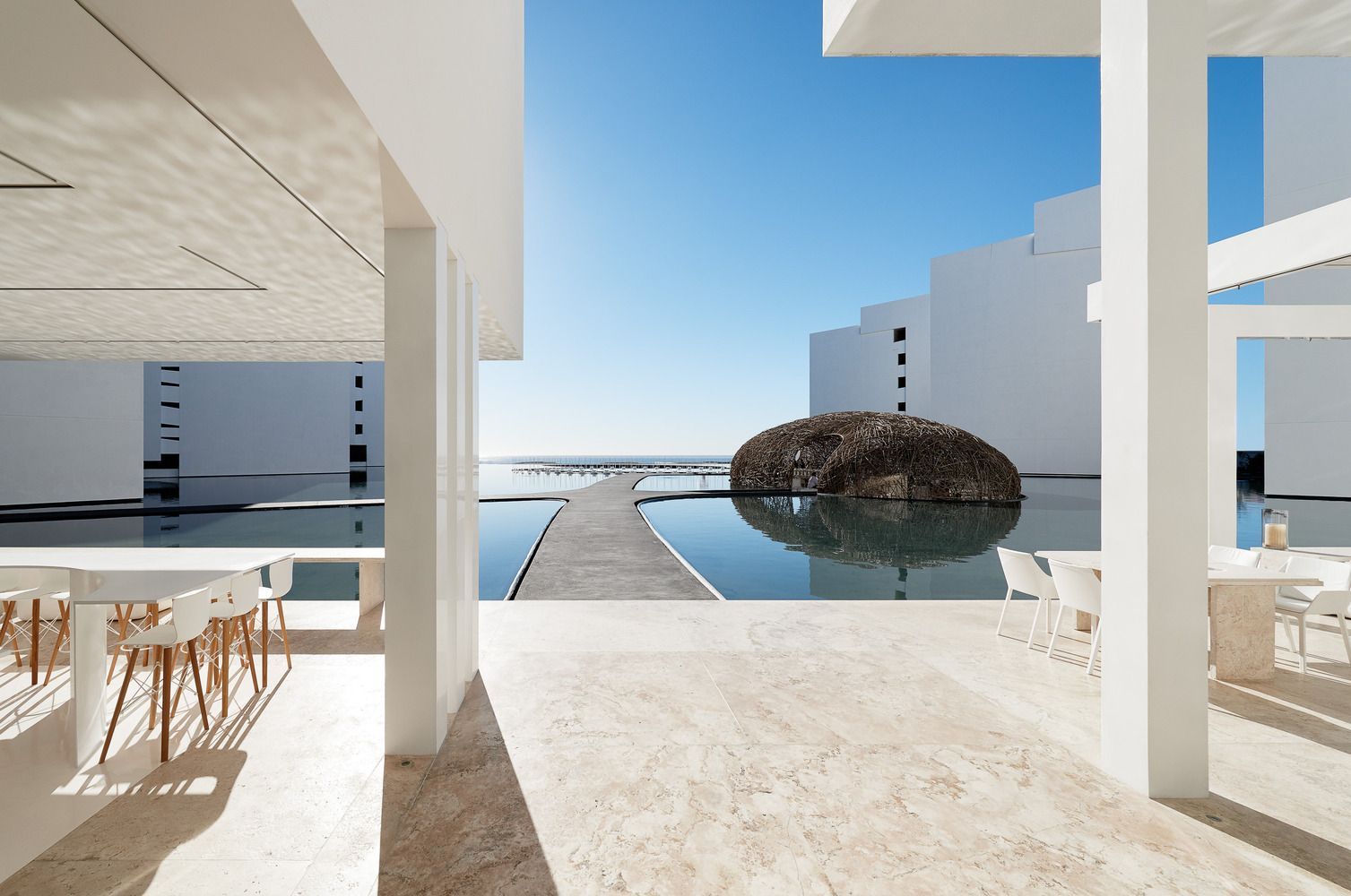 Patio blanco moderno con vistas a una piscina y al océano bajo un cielo azul brillante.