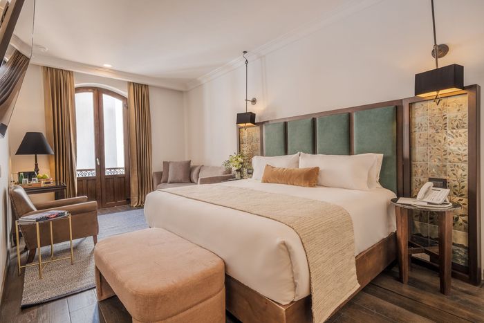 Elegante dormitorio con cama king size, cabecero color turquesa, iluminación cálida y zona de estar junto a la ventana.