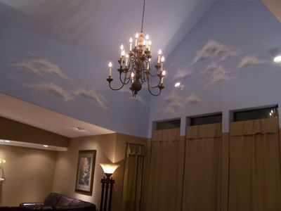A chandelier hangs from a vaulted ceiling painted light blue with clouds, above a room with tan walls and tall drapes.