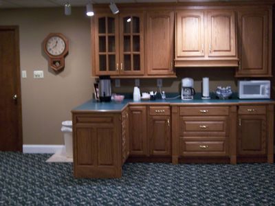 A kitchenette with wood cabinets, green countertops, and a coffee maker, located next to a wall clock in a carpeted room.