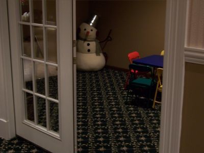 A large snowman decoration in the corner of a dimly lit room with a children's table and small colorful chairs.