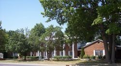 A two-story brick house with white pillars and a green lawn, shaded by mature trees under a clear blue sky.