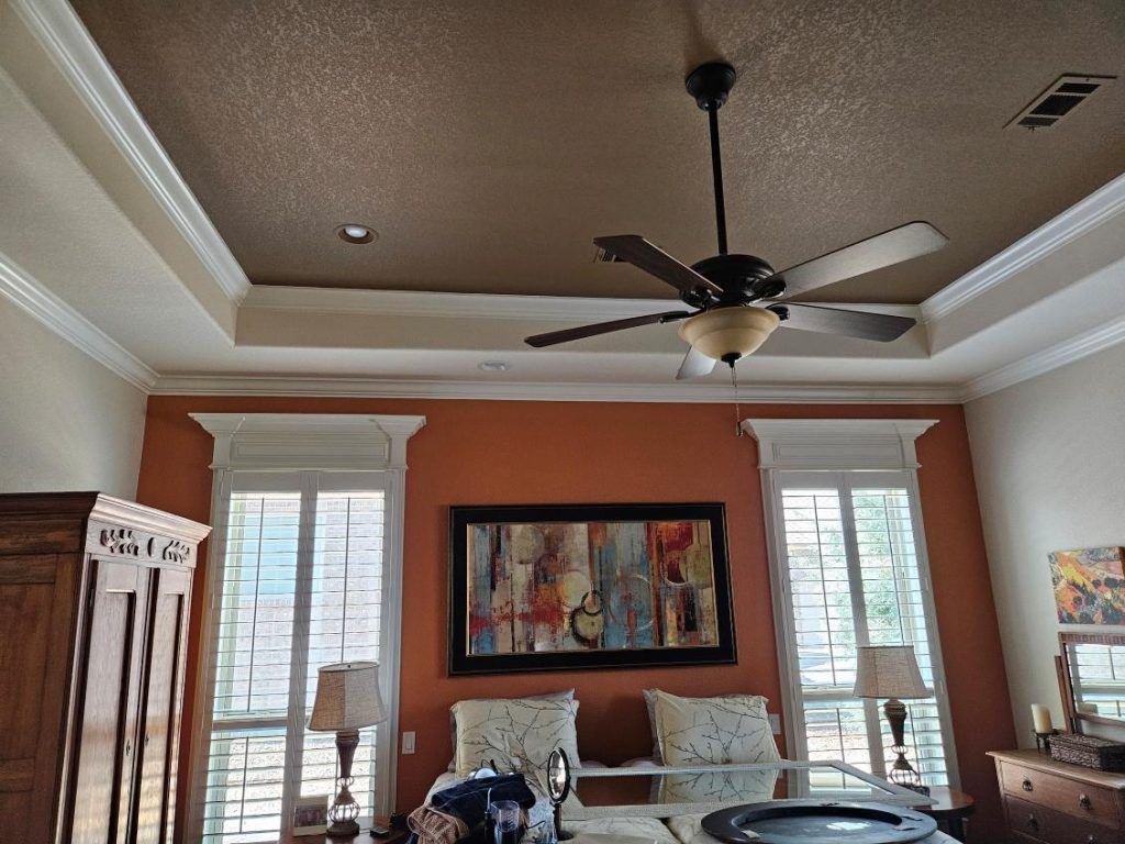 A room with an orange wall, crown molding, ceiling fan, windows, and a painting.