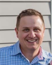 Smiling man in a blue checkered shirt standing against gray siding