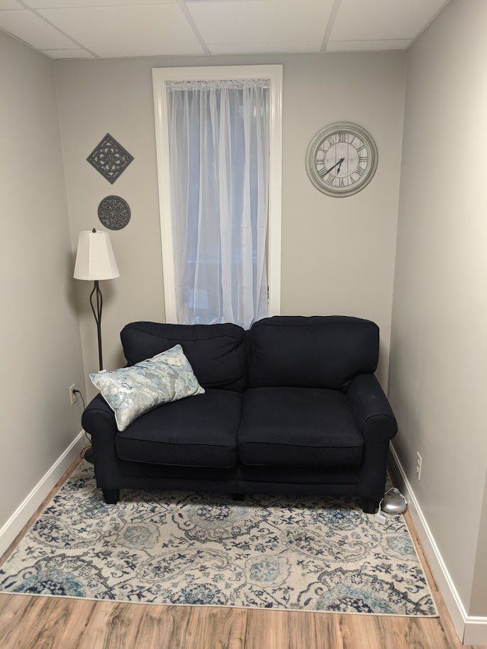 Small living room with navy loveseat, patterned rug, lamp, wall clock, and window with sheer curtain