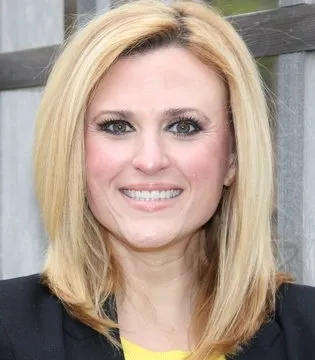 Smiling woman with blonde hair in a black jacket, standing outdoors against a wooden fence.