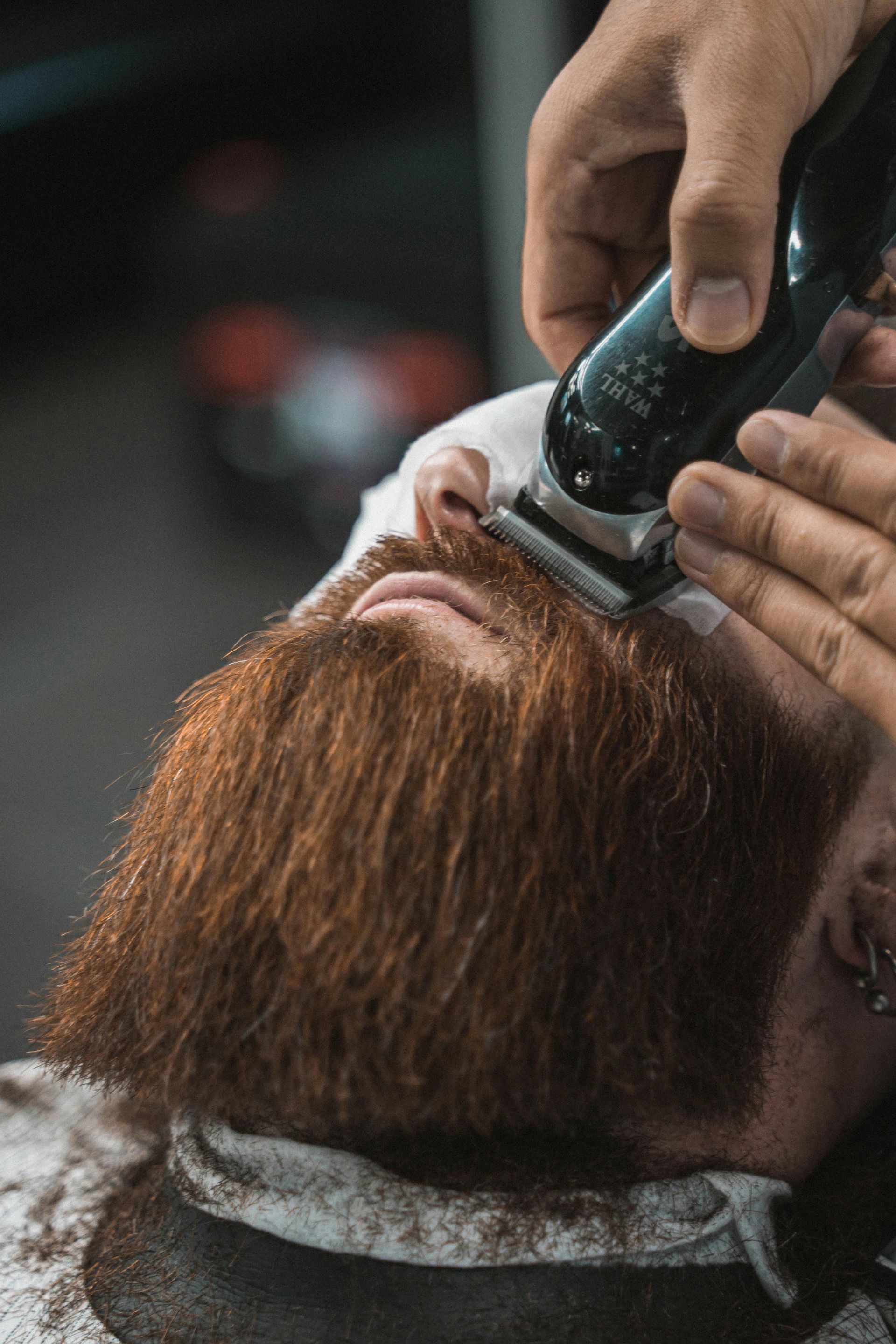 A barber uses electric clippers to trim a man's thick, reddish-brown beard.
