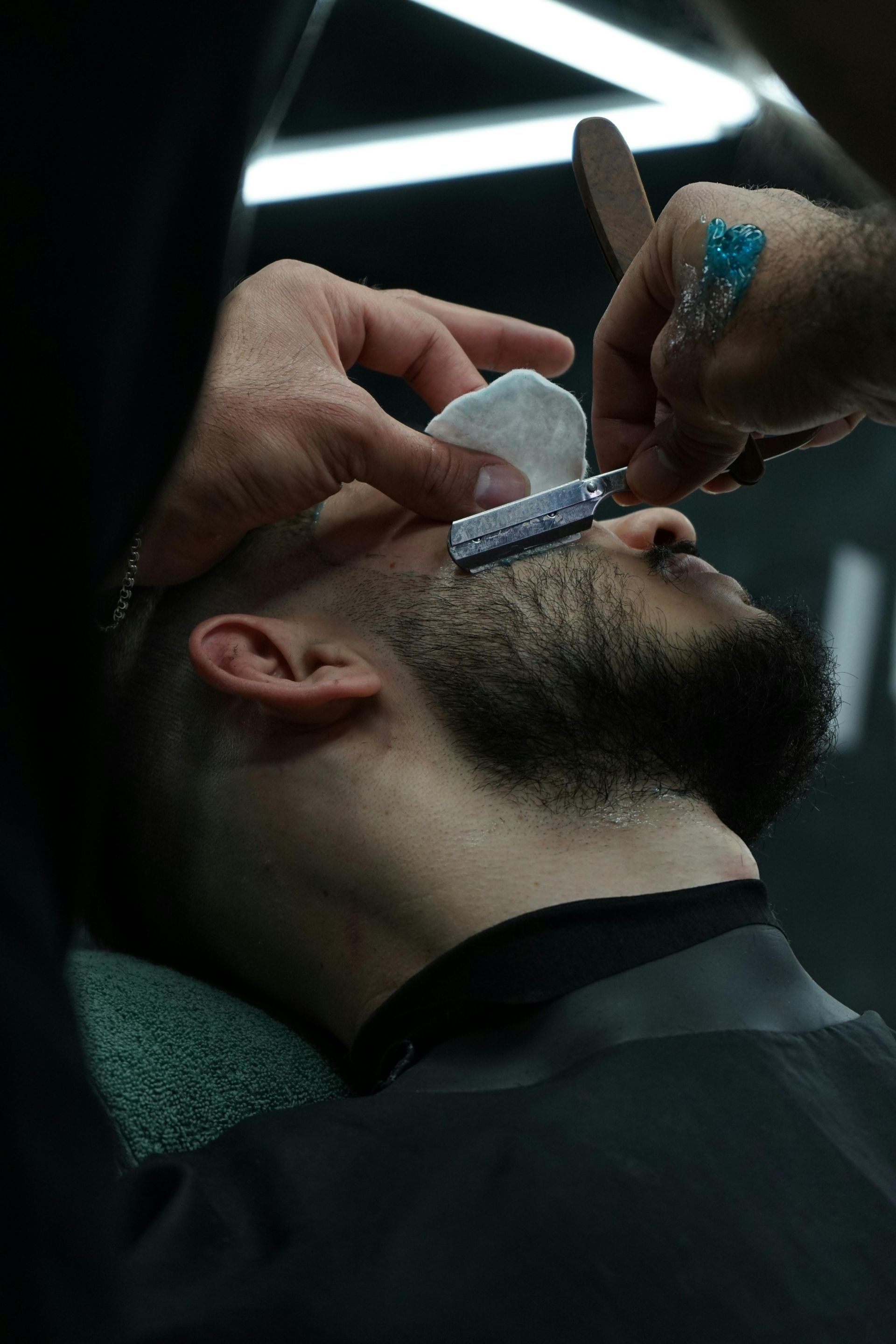 A barber carefully uses a straight razor to shave a customer's cheek in a dimly lit, professional salon setting.