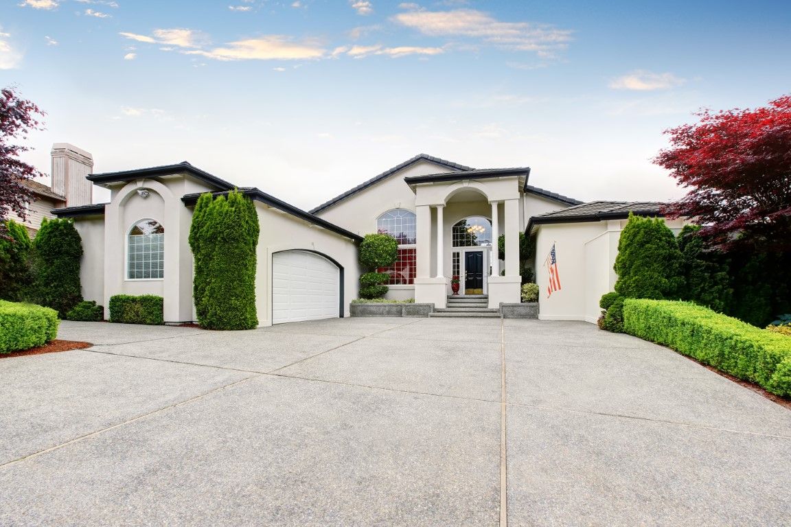Large modern house with white stucco exterior, arched entry, and wide driveway under a blue sky