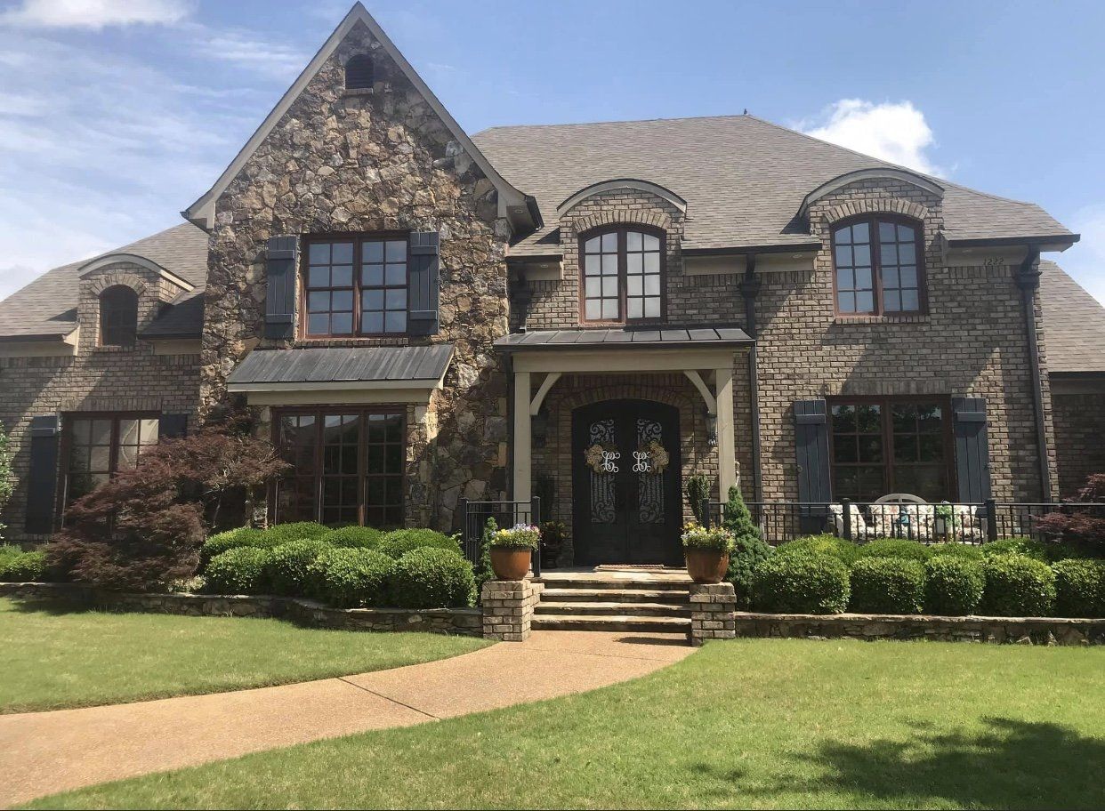 Stone house with a brown roof, shutters, and walkway on a sunny day.