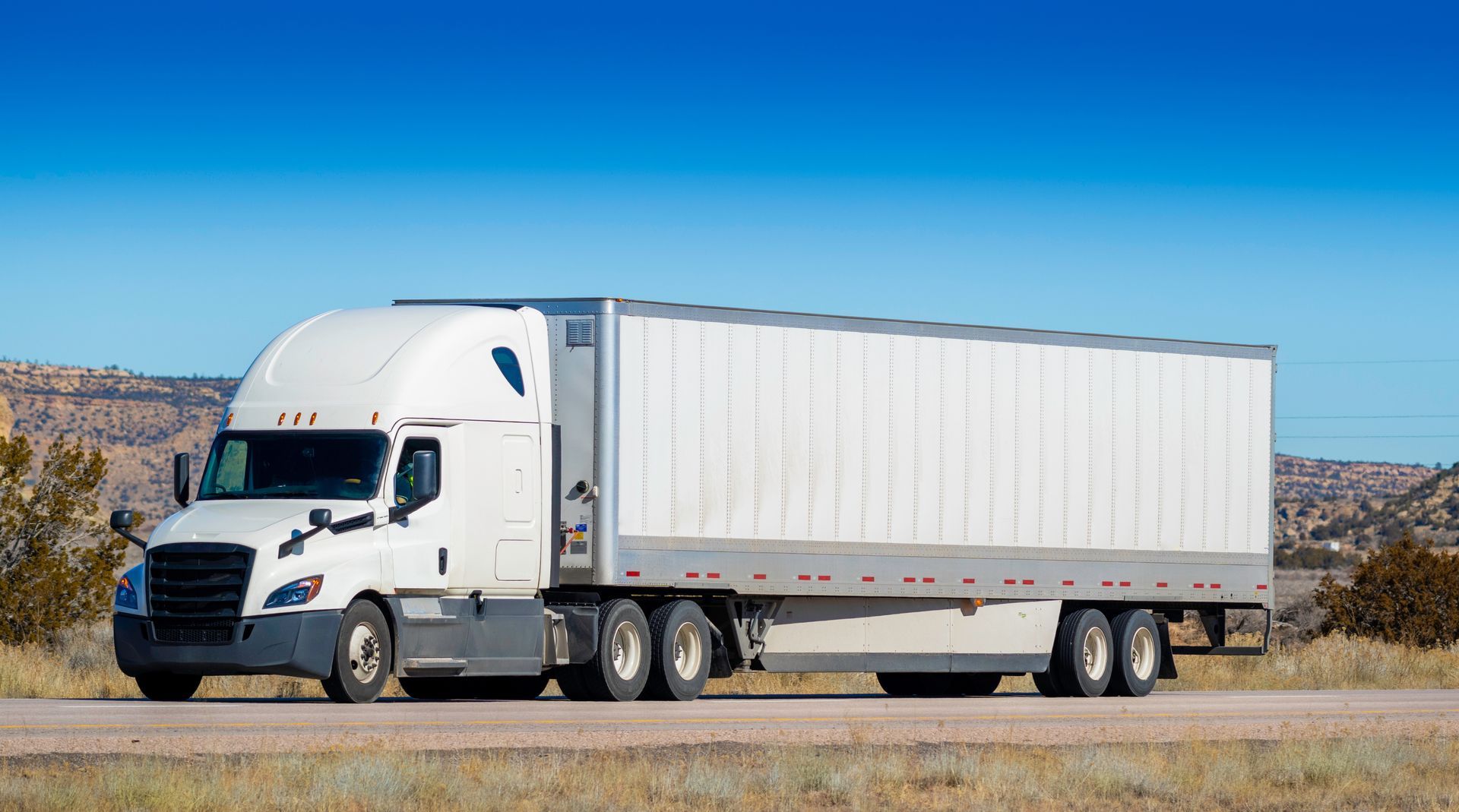 White semi-truck with a box trailer driving on a road under a blue sky.