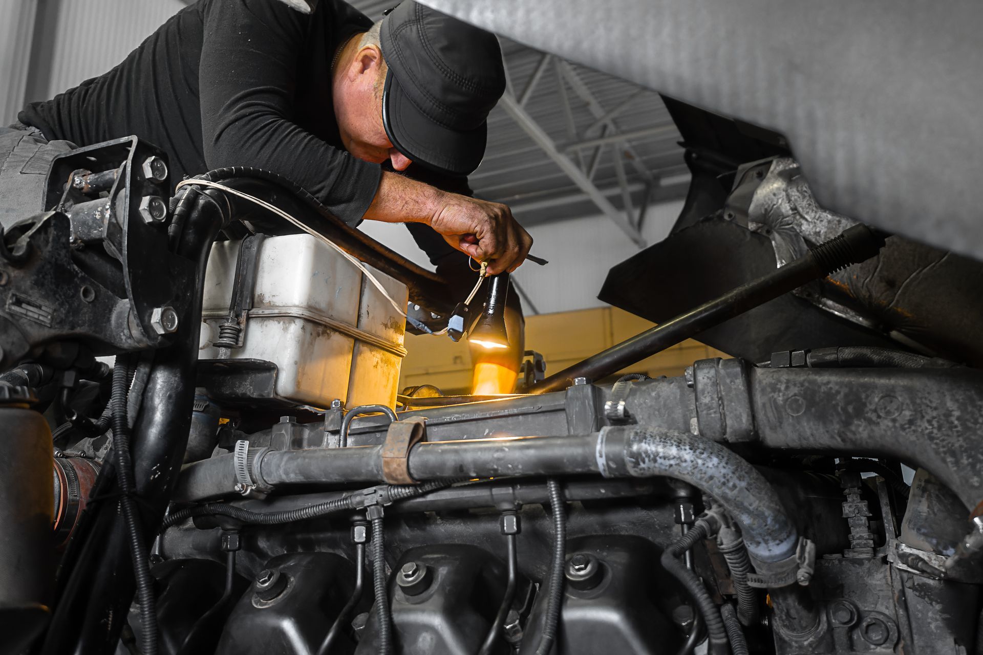 Mechanic leaning over a truck engine, using a tool under bright workshop lights.