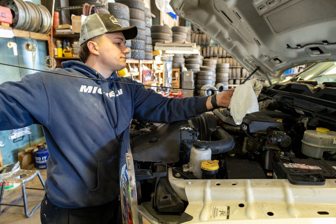 Mechanic in blue hoodie working under an open car hood in a cluttered auto shop