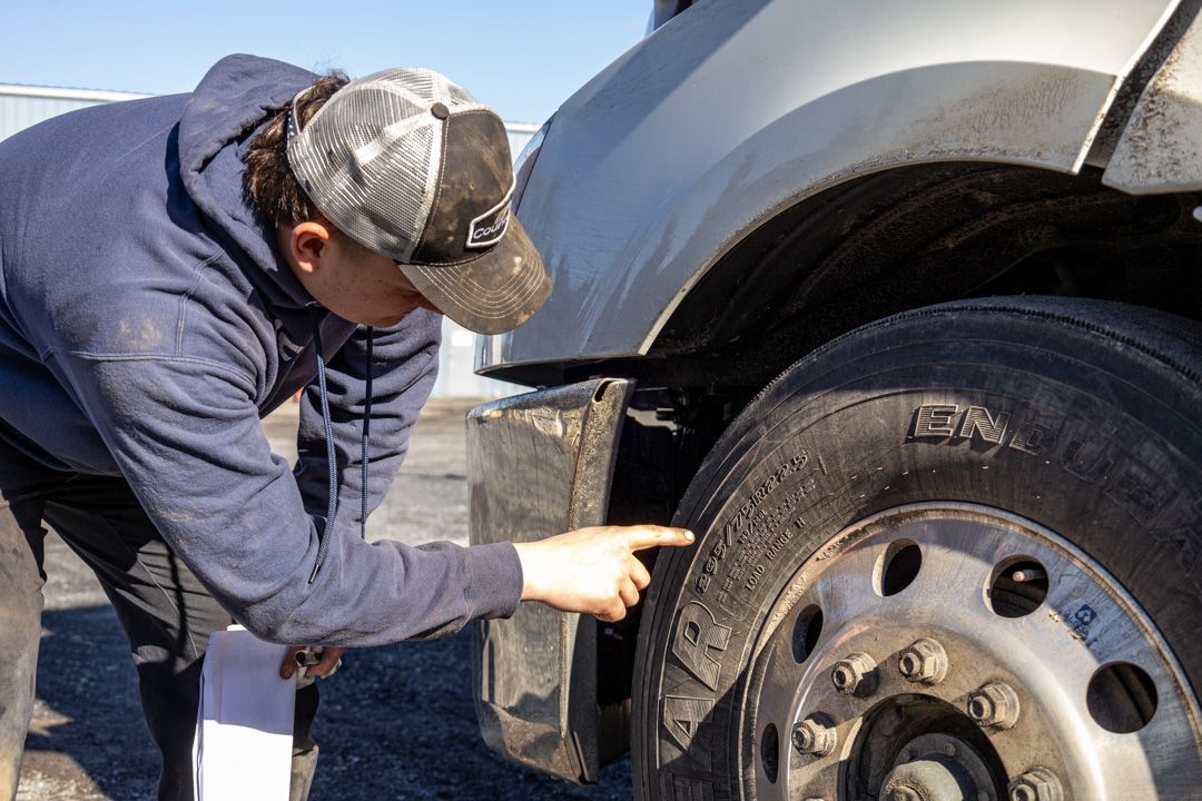Person inspecting a truck tire and wheel while crouching beside the vehicle outdoors.