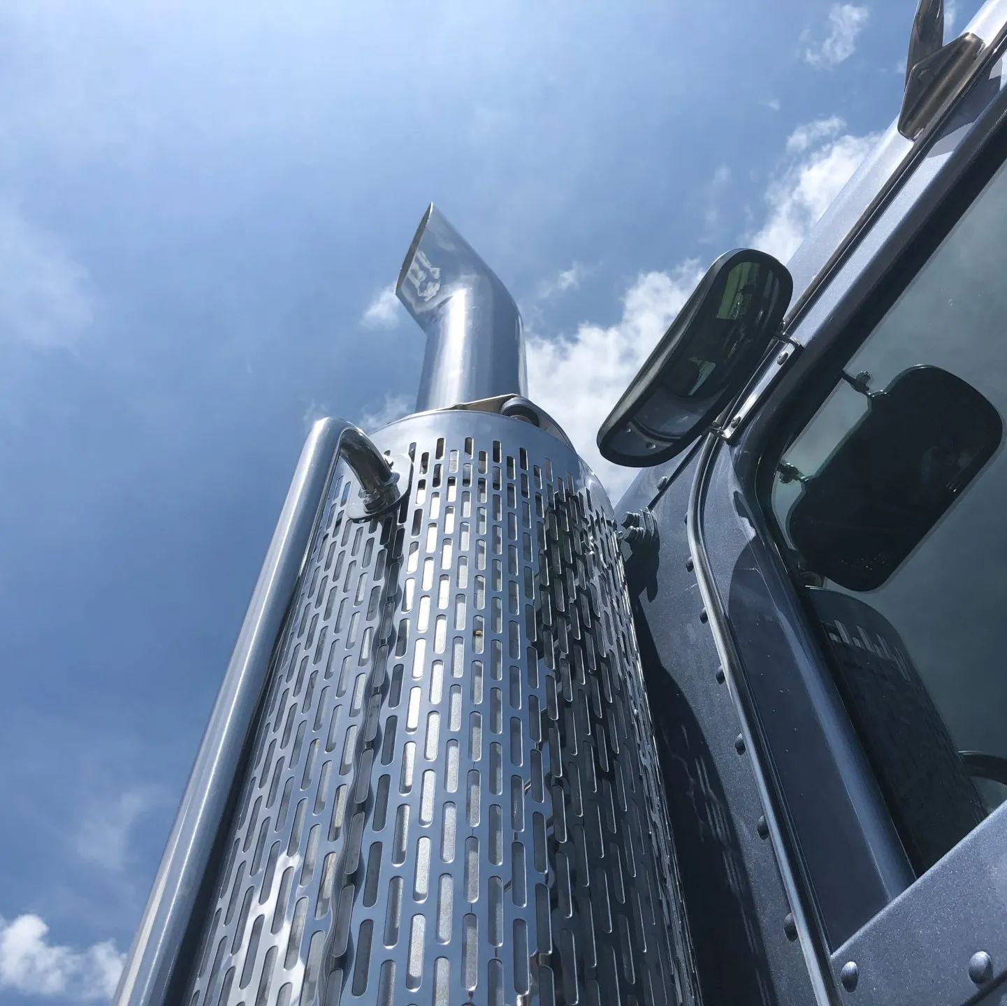 Chrome truck exhaust stack against blue sky, viewed from below beside a truck cab.