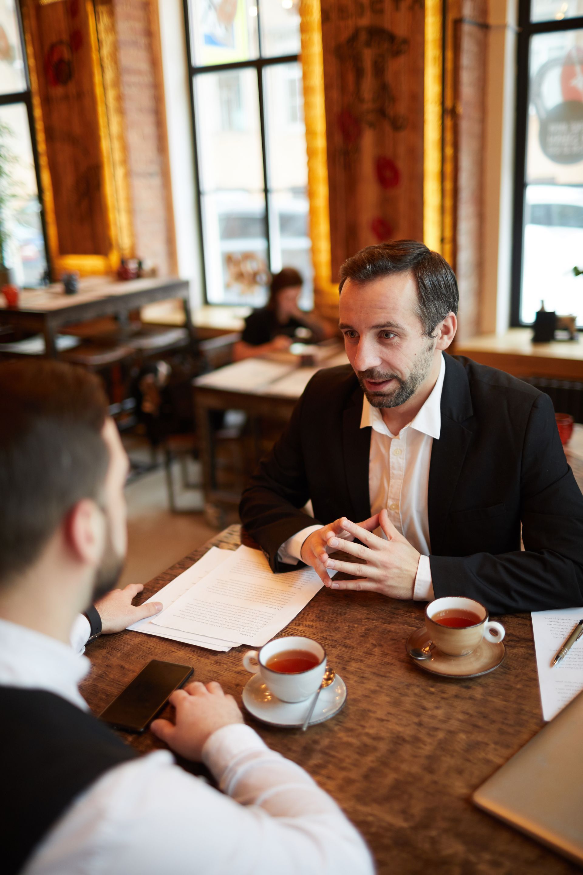 Two people talking at a café table with coffee cups and papers on a wooden table.
