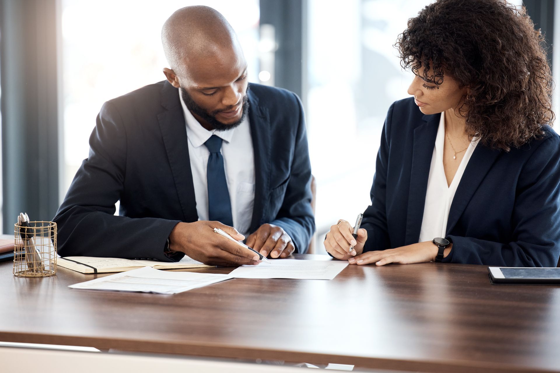 Two professionals in dark suits review documents at a desk in a bright office