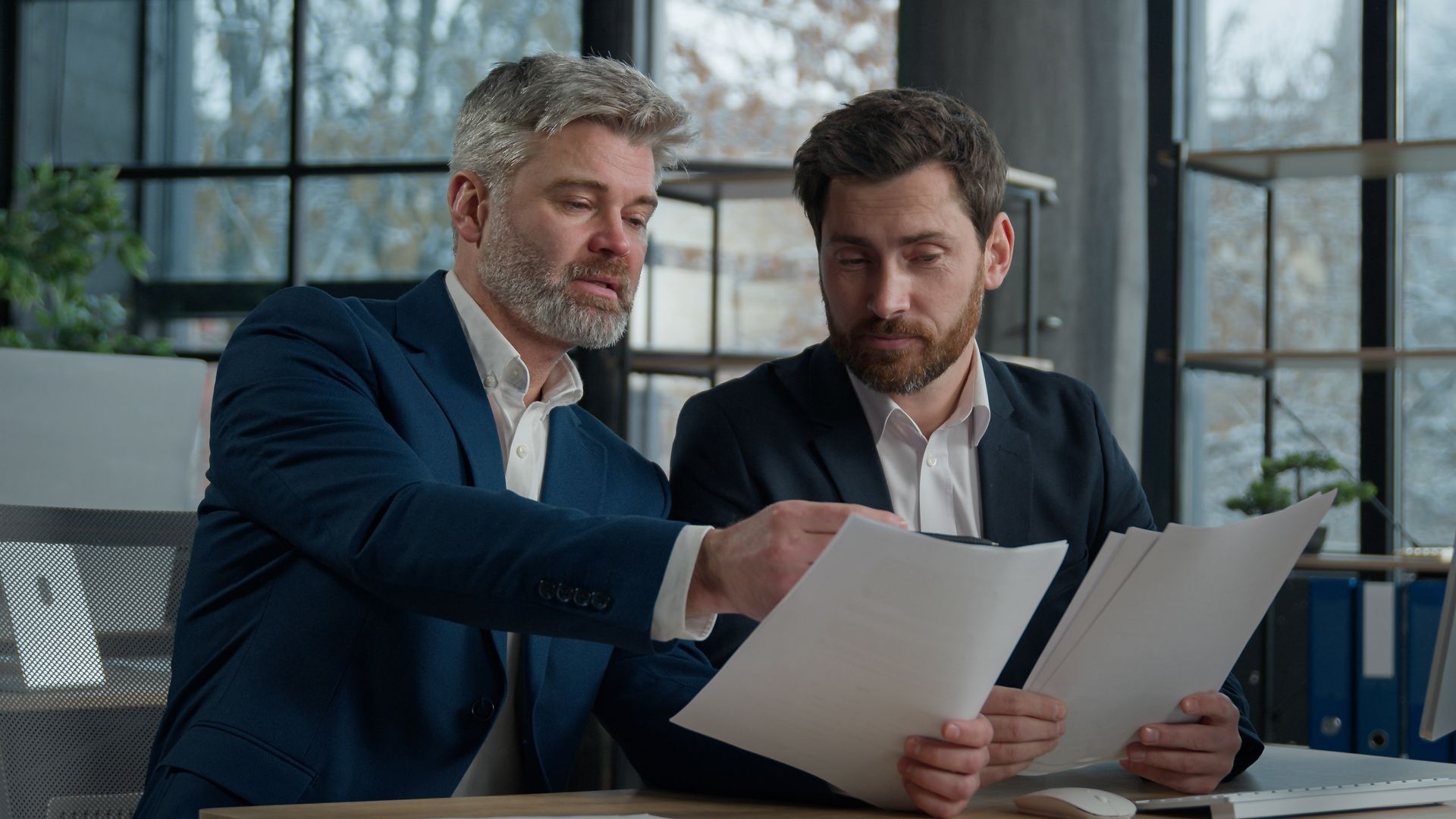 Two men in suits review documents at a desk in a modern office
