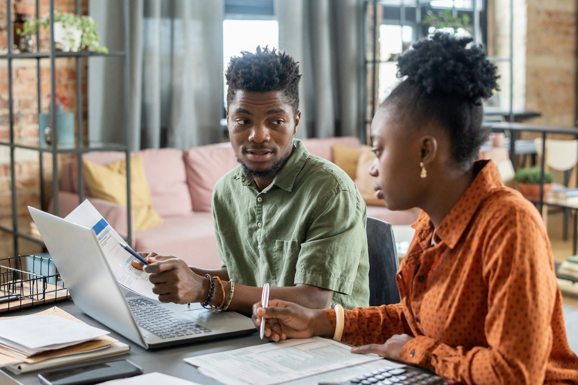 Two coworkers discussing a laptop at a desk in a bright office, with papers and coffee nearby.