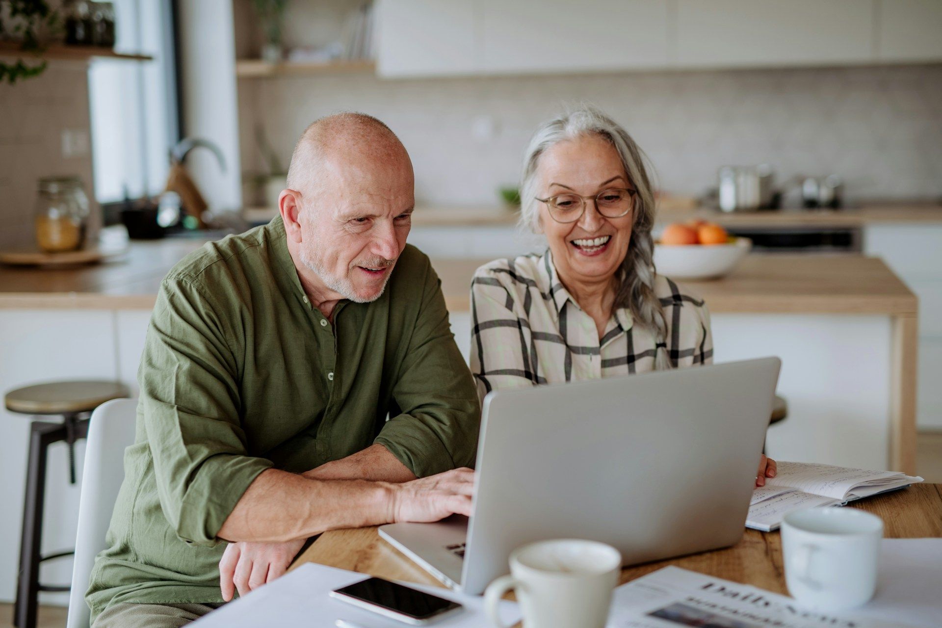 Two people smiling at a laptop in a bright kitchen, with coffee cups on the table.