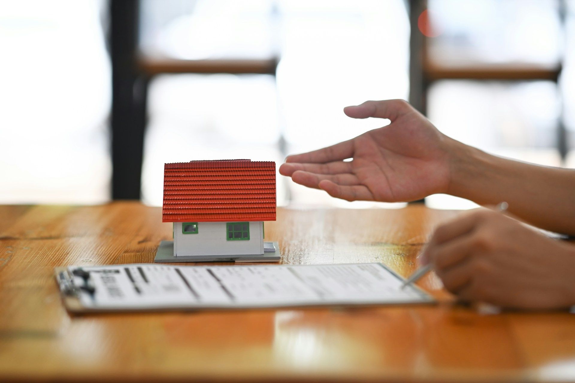 Hand pointing at a small house model beside a document on a wooden table
