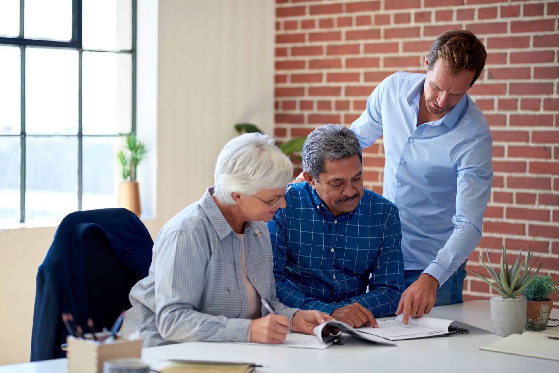 Three coworkers review documents at a desk in a bright office, one standing and pointing.