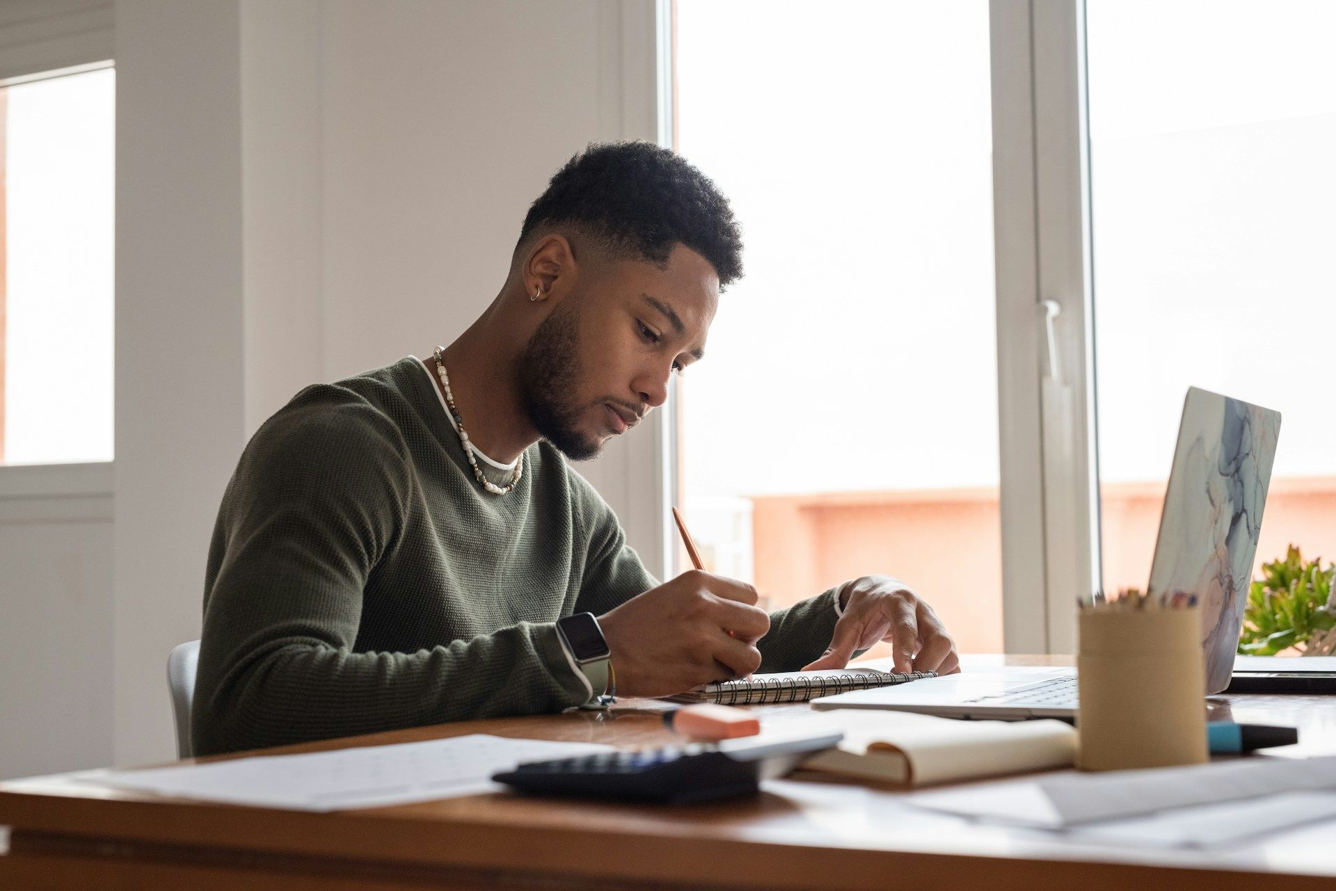 Person sketching at a desk beside an open notebook and coffee mug in a bright room