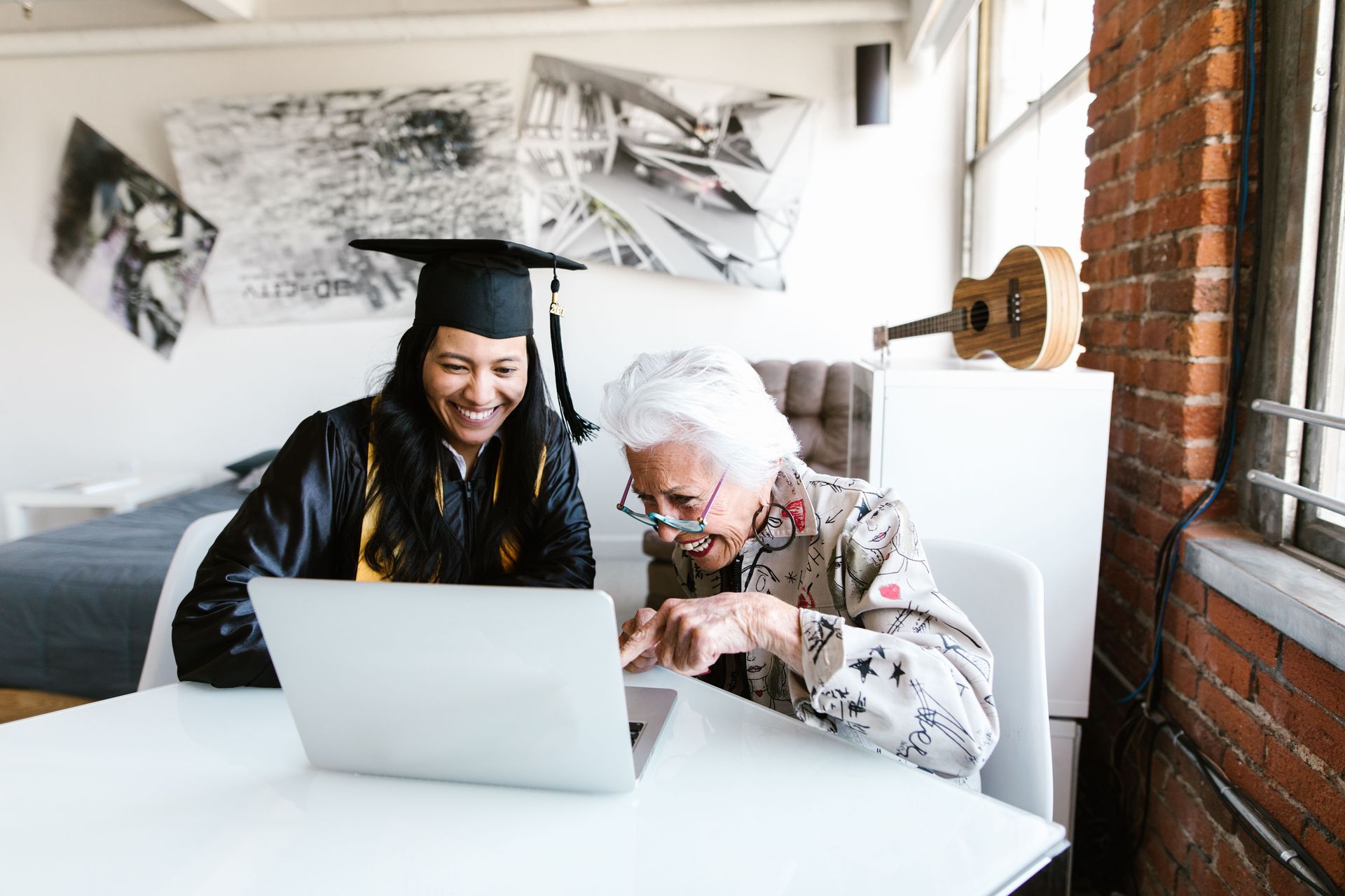 Two people smiling at a laptop in a bright room, one wearing a graduation cap