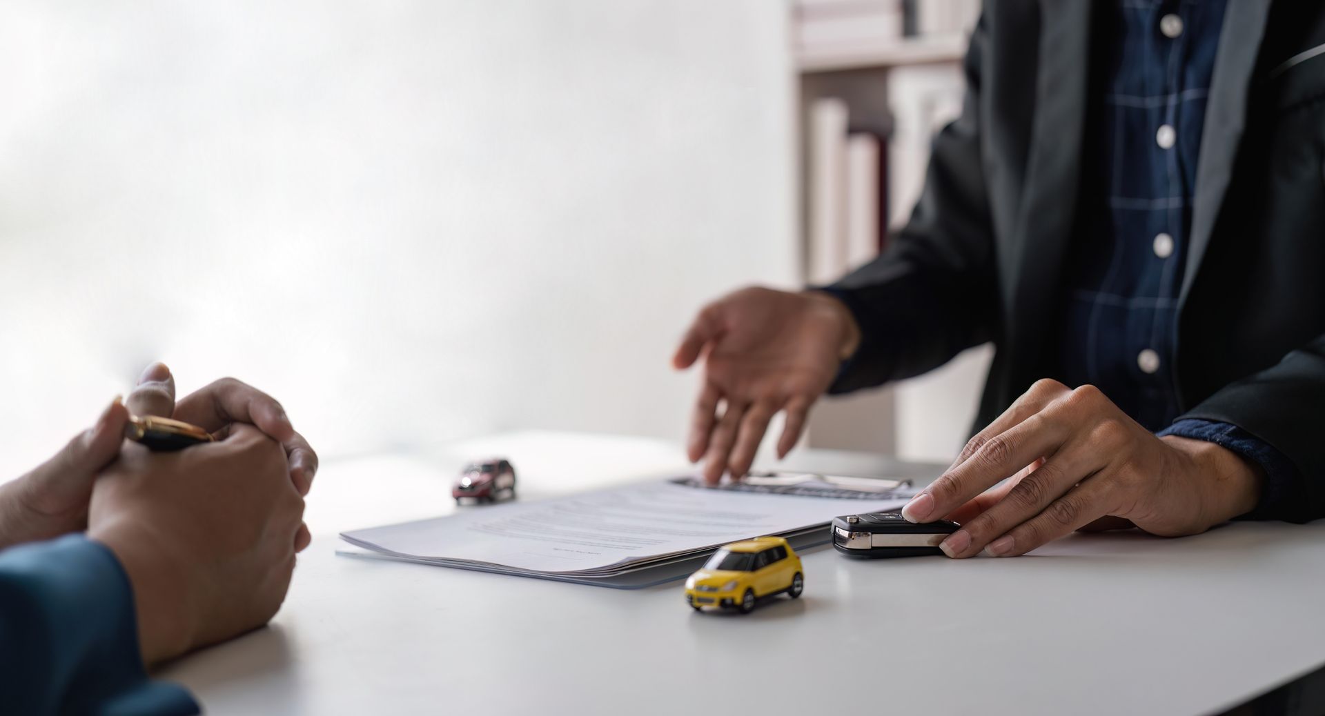 Hands over a document on a white table beside two toy cars during a meeting.
