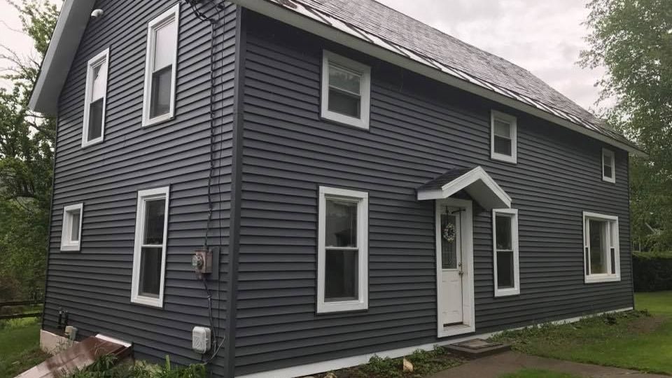 a large gray house with white trim and windows is sitting on top of a lush green field .