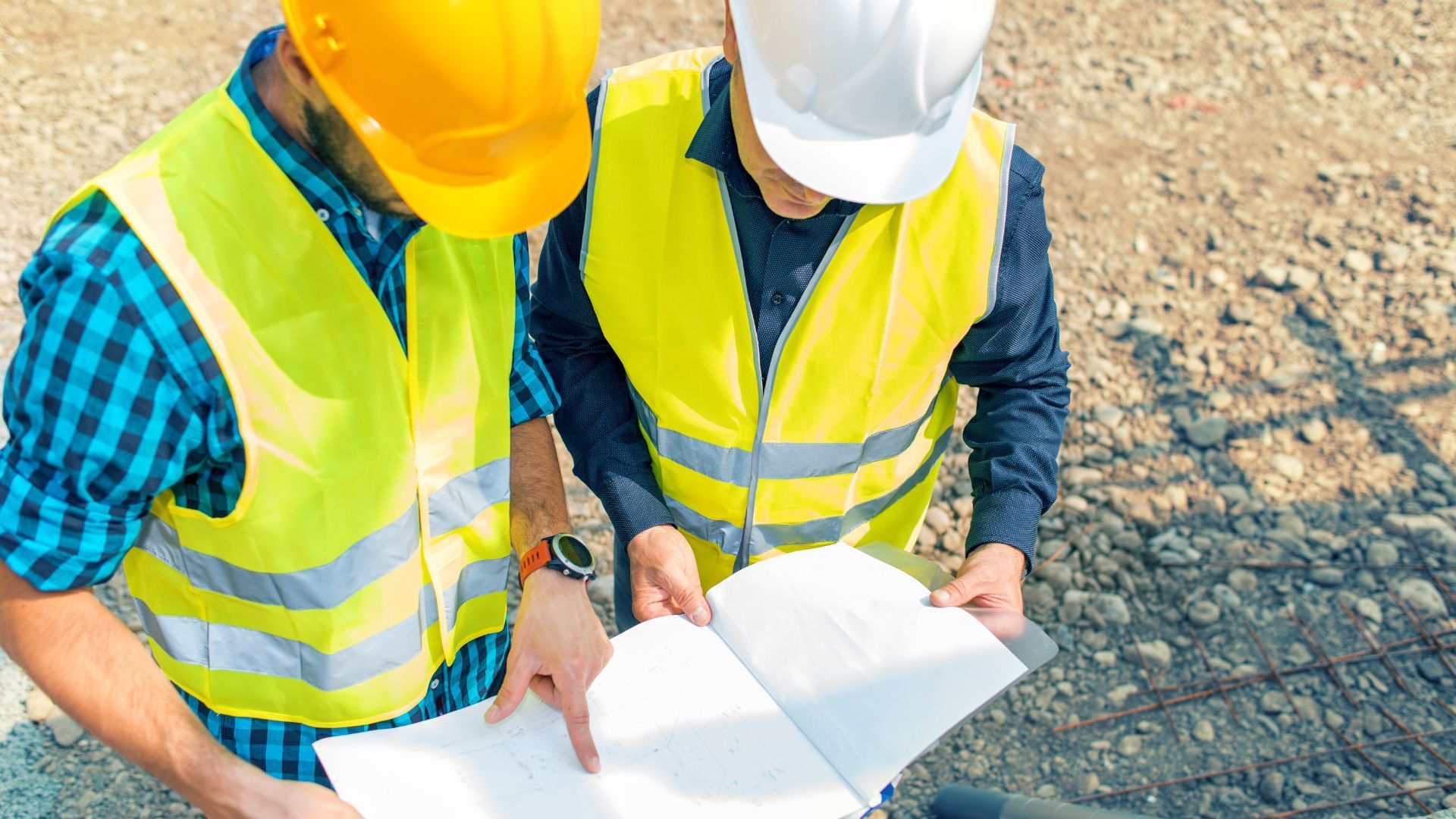 Deux ouvriers du bâtiment, vêtus de gilets haute visibilité et de casques de chantier, examinent des plans sur un chantier.