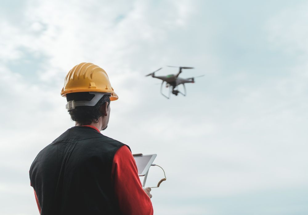 Une personne portant un casque et un gilet jaunes pilote un drone à l'aide d'une tablette sur fond de ciel nuageux.