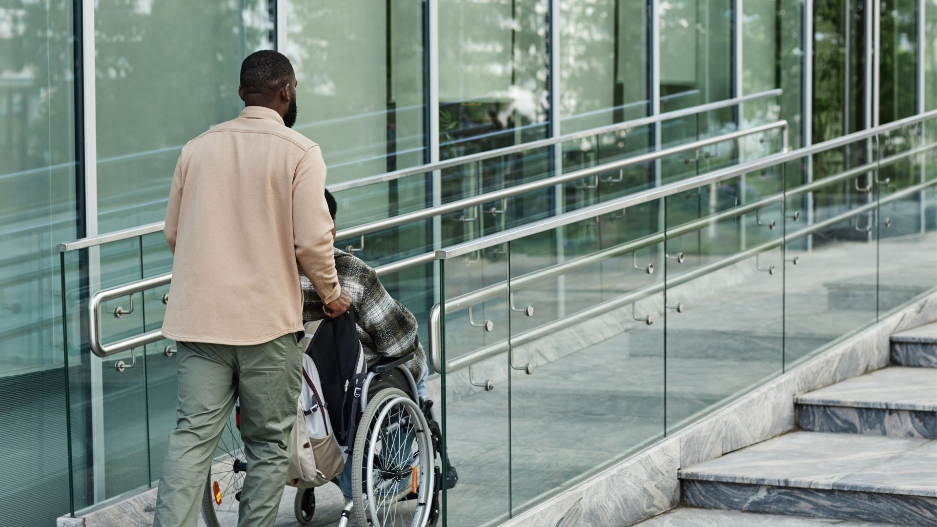 Une personne pousse une autre personne en fauteuil roulant sur une rampe longeant un bâtiment aux murs de verre et aux escaliers.