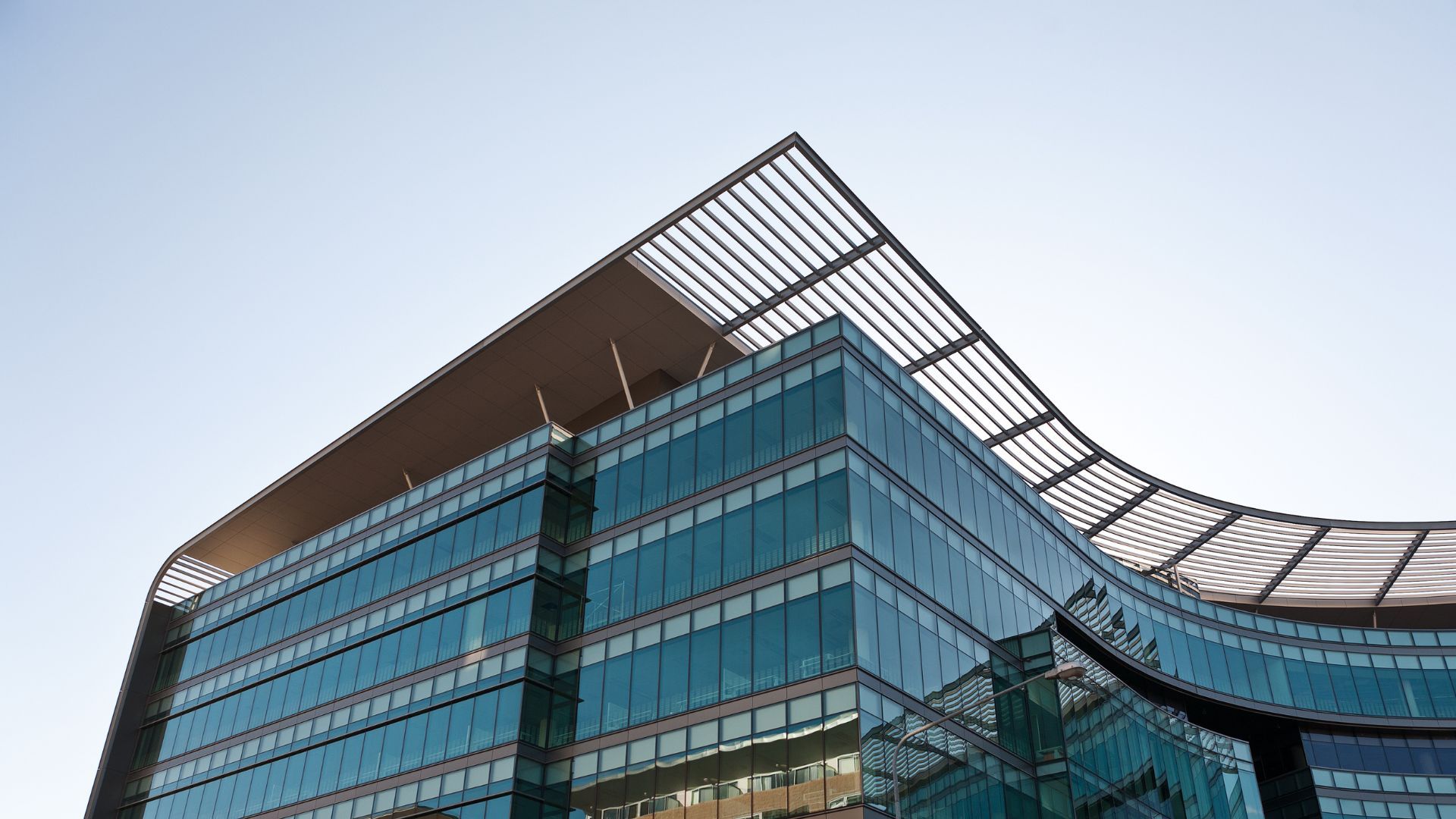 Vue en contre-plongée d'un immeuble de bureaux moderne en verre, avec une toiture incurvée à lames, se détachant sur un ciel bleu clair.