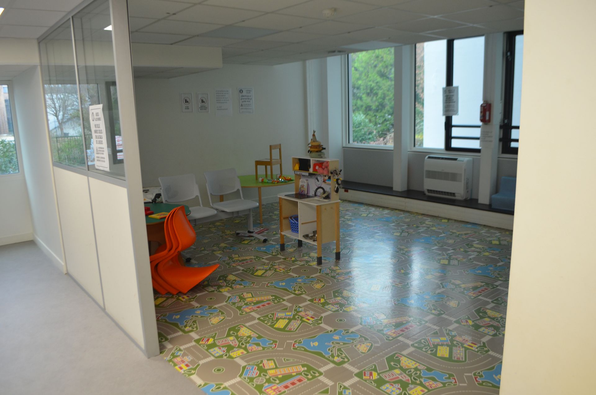 Un espace de jeux pour enfants avec un tapis à motif de carte routière, des chaises à leur taille et une petite étagère à jouets en bois.