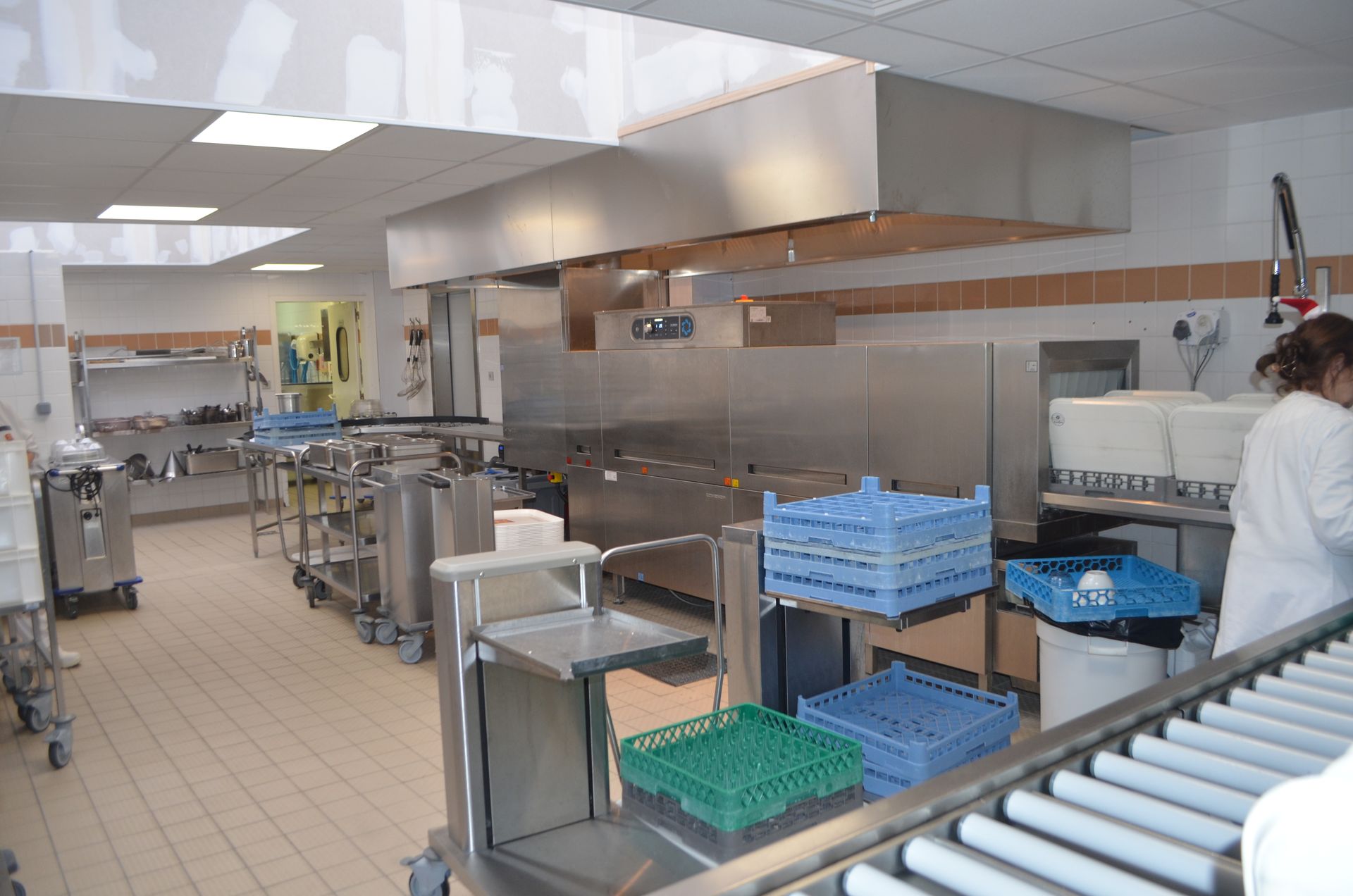 Une zone de lavage de lave-vaisselle dans une cuisine commerciale comprenant des équipements en acier inoxydable, des convoyeurs à rouleaux et des piles de caisses en plastique.