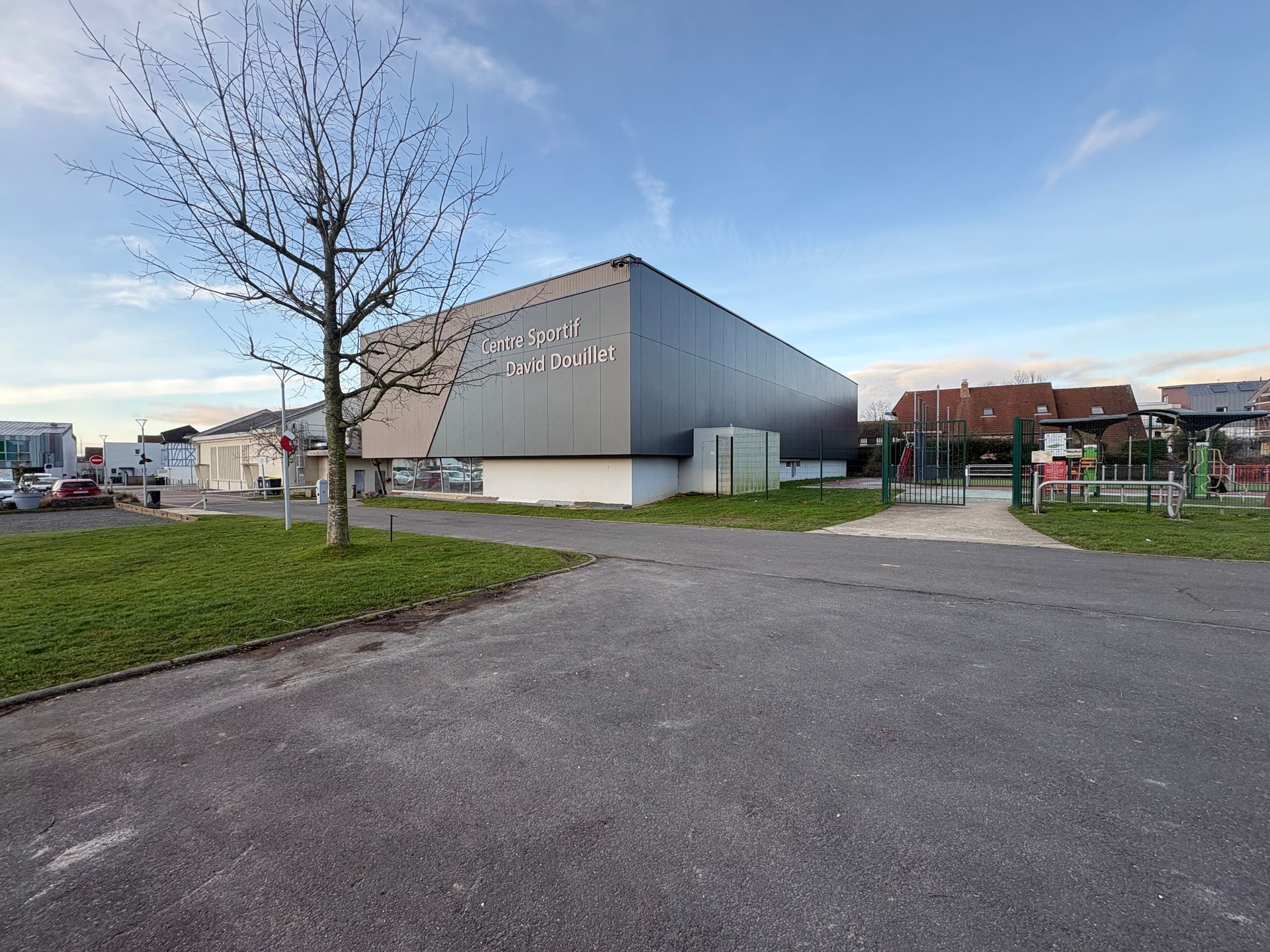 Un bâtiment industriel gris, moderne et rectangulaire, se dresse sous un ciel lumineux, avec un arbre dénudé au premier plan.