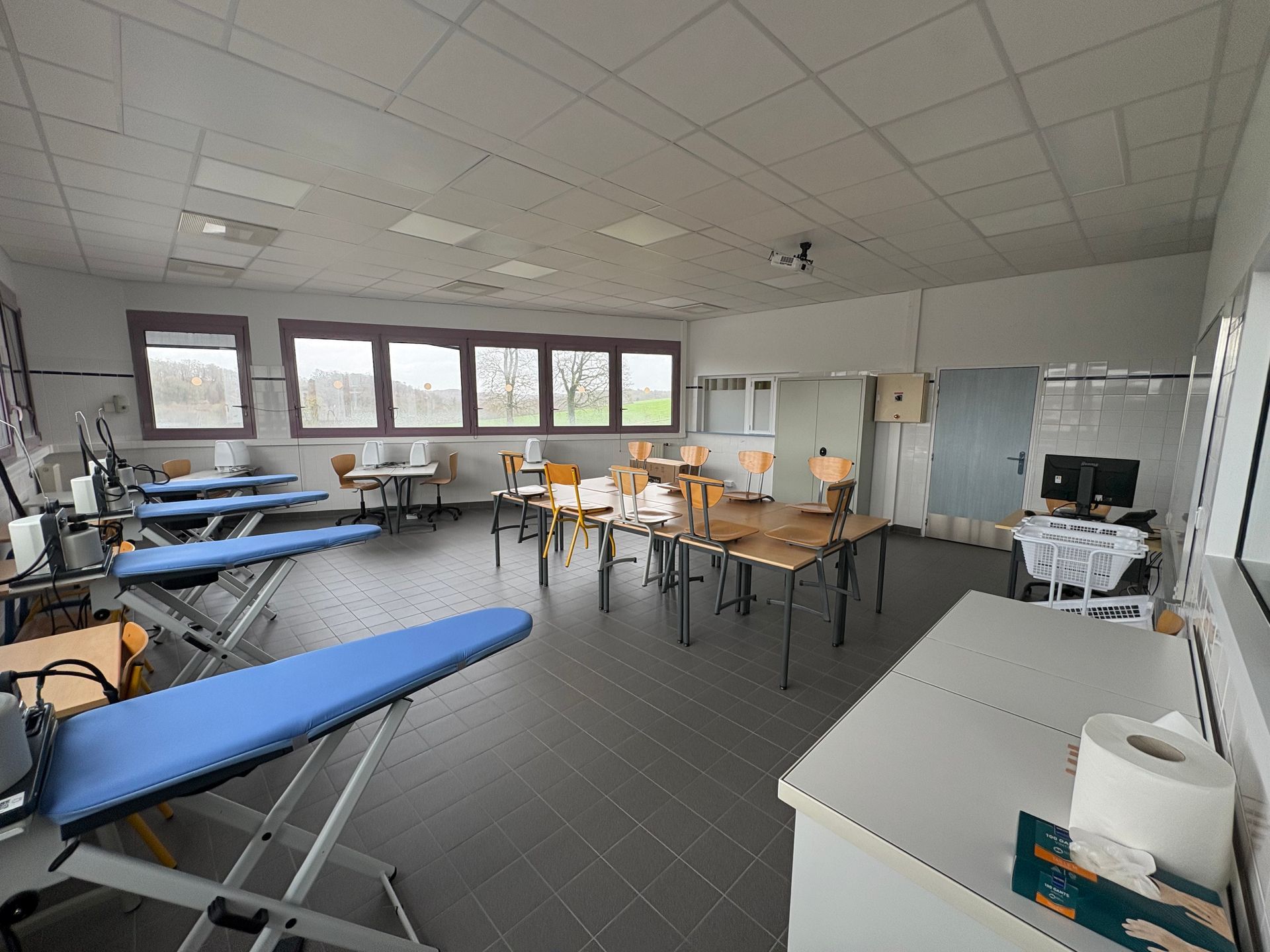 Une salle de classe ou de formation vide, avec des planches à repasser bleues, des tables en bois, des chaises et de grandes fenêtres.