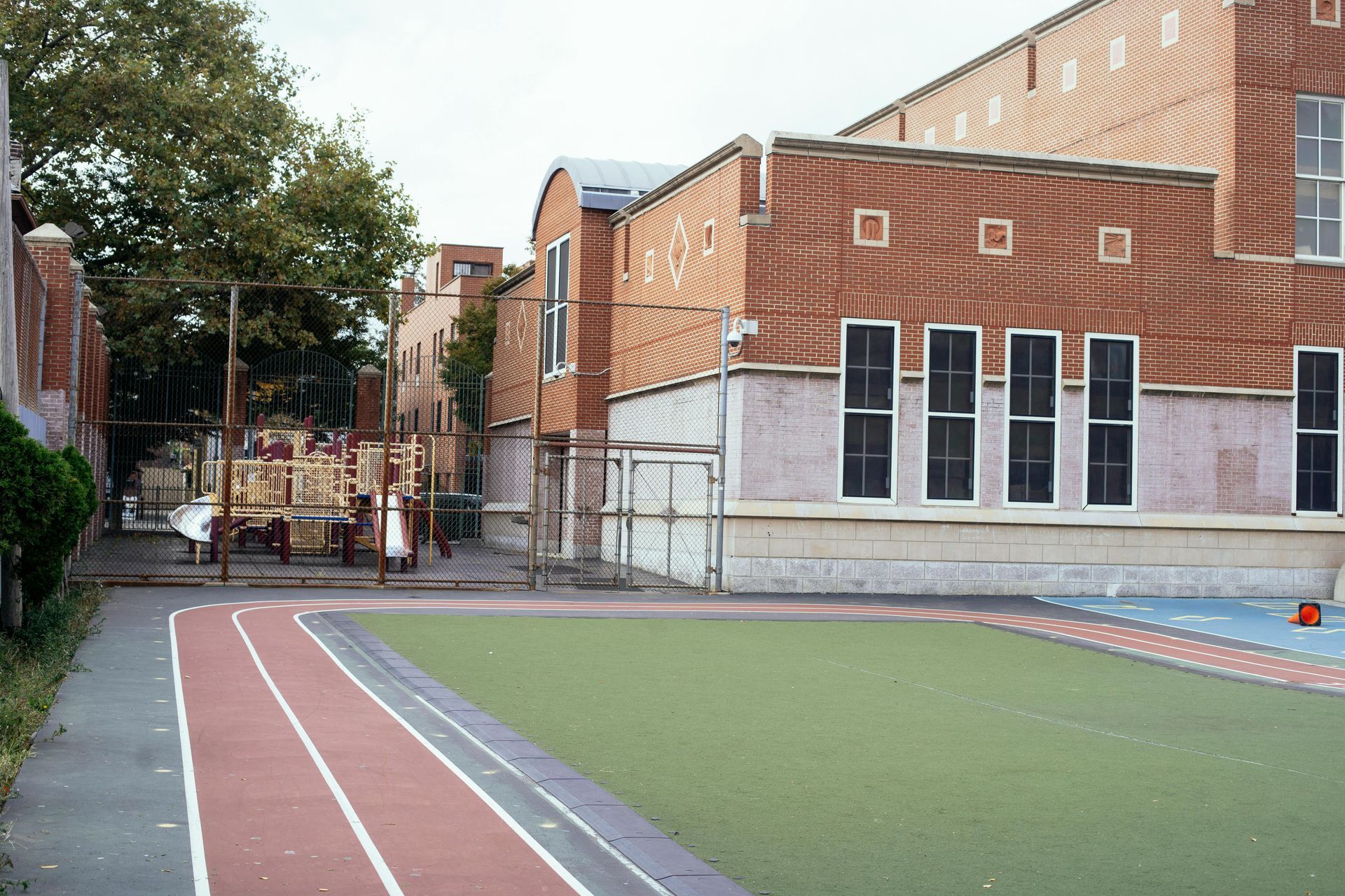 Une aire de jeux avec une piste de course rouge et un terrain vert jouxtant un bâtiment scolaire en briques, sous un ciel nuageux.