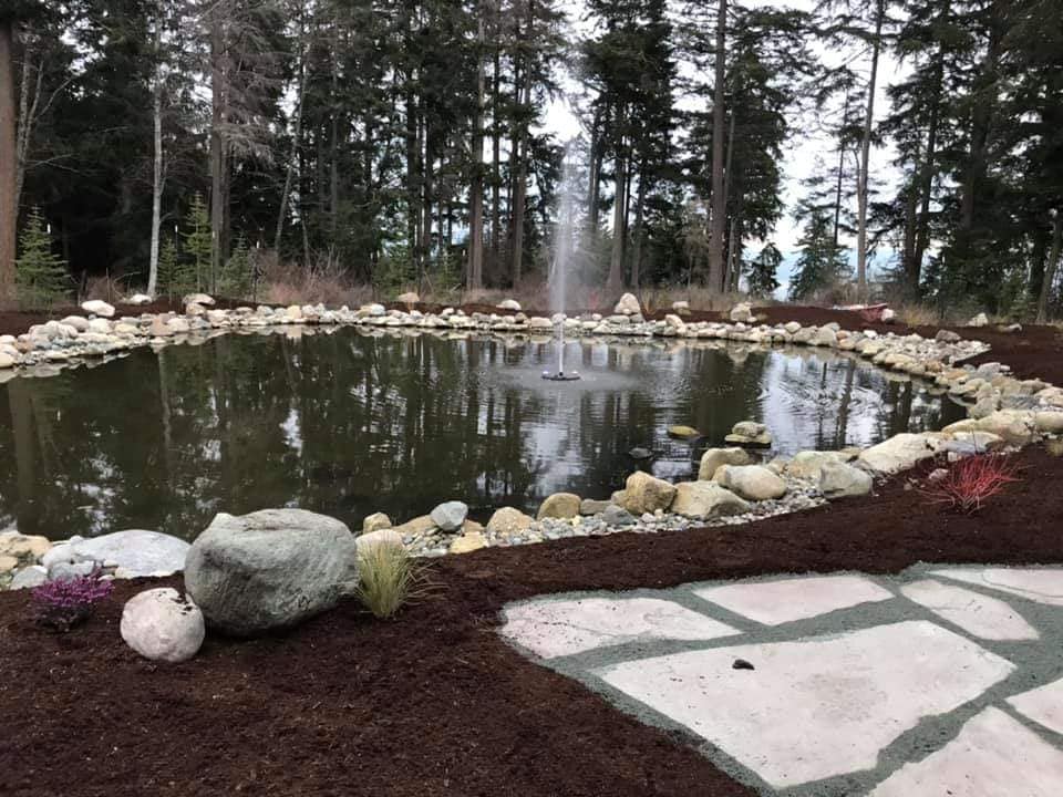 Pond with fountain in a forest setting, surrounded by rocks and a stone patio.