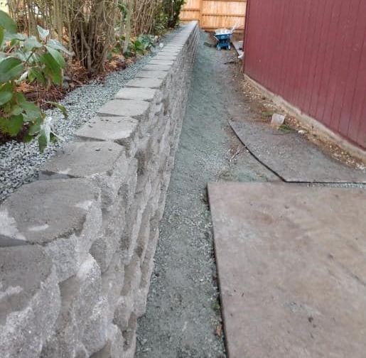 Stone retaining wall next to gravel and concrete pathway, bordering a building and greenery.