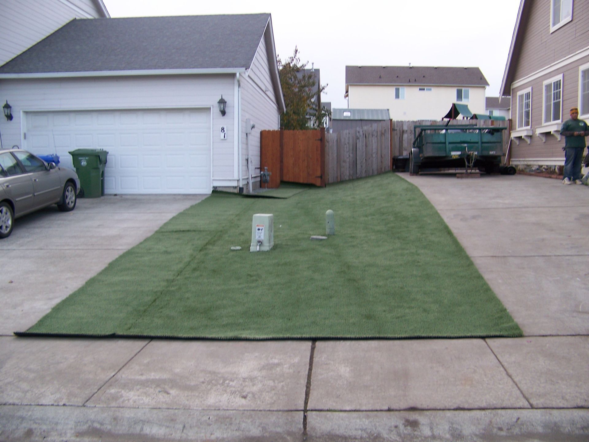 Green artificial turf covers a driveway section, with a car, garage, and dumpster visible.