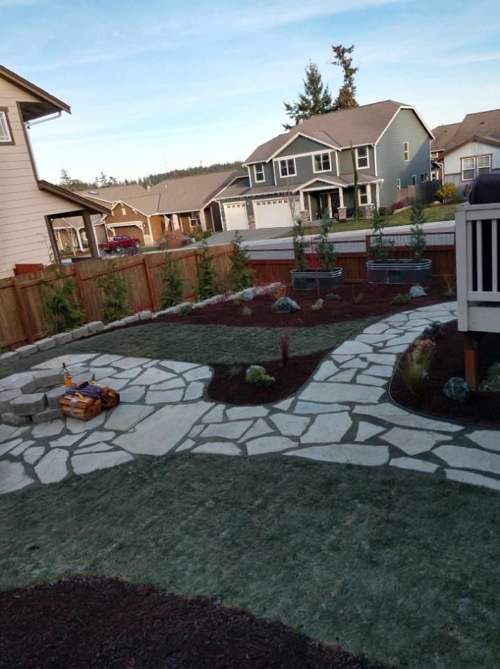 Backyard with stone pathway, fire pit, and landscaping. Houses and blue sky in the background.