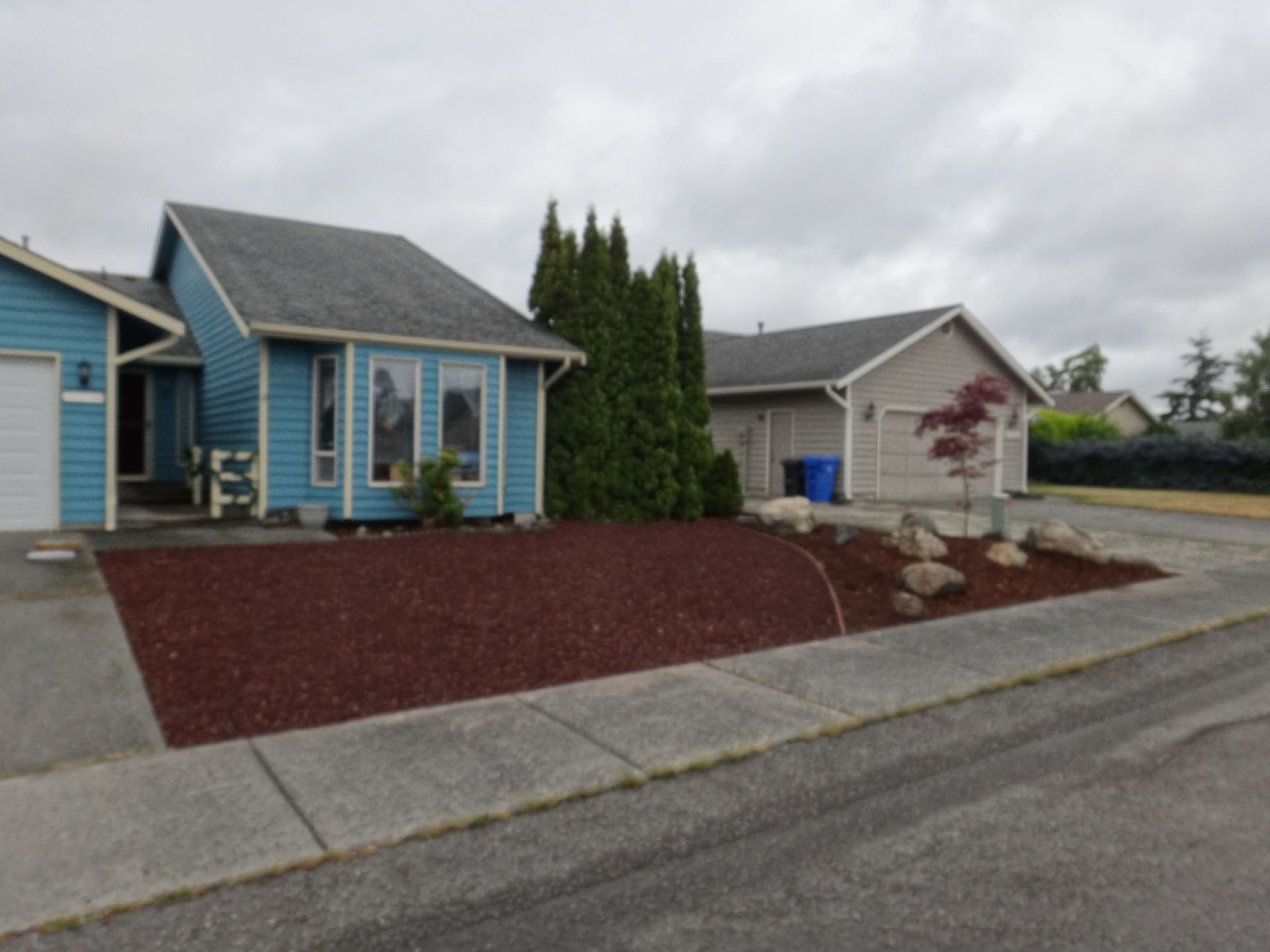 Blue house with detached gray garage, red mulch landscaping, cloudy sky.