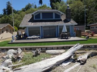 Blue waterfront cottage with large windows, deck, and driftwood on the beach.