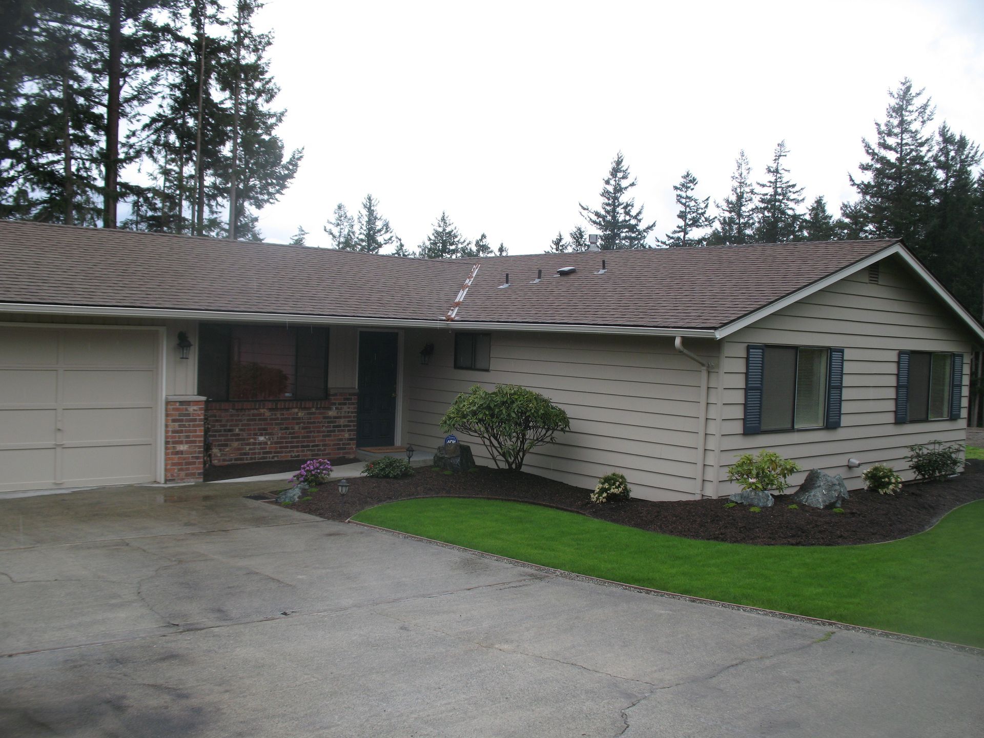 Tan ranch-style house with a garage, brown roof, and dark blue shutters, set against a backdrop of trees.