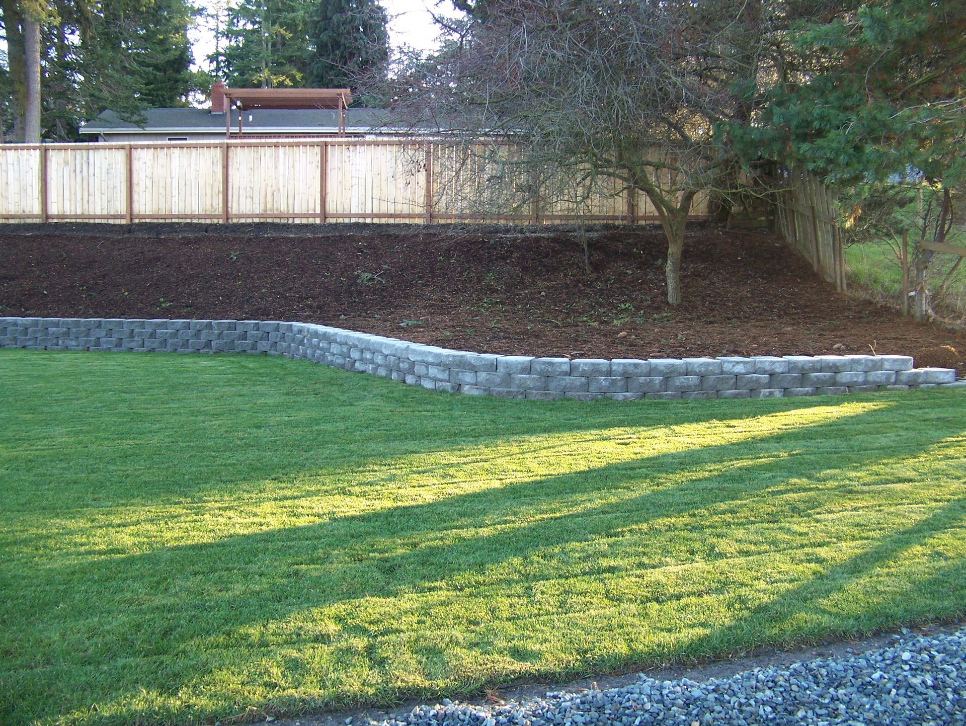 Grassy yard with a retaining wall, mulch, and a wooden fence; trees and a house in the background.