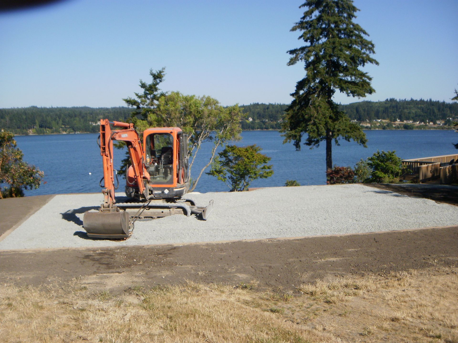 Orange excavator on a gravel pad overlooking a lake with trees.