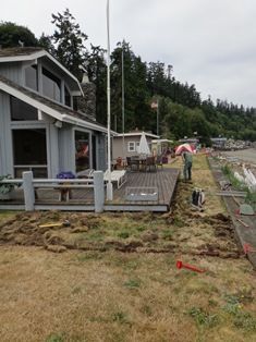 A person rakes grass on a lawn next to a waterfront building with a deck on a cloudy day.